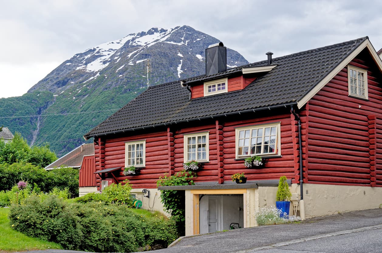 Rødt laftehus med svart tegltak og fjell i bakgrunnen, brukt som illustrasjon i artikkel om å bruke megler eller advokat ved boligsalg.