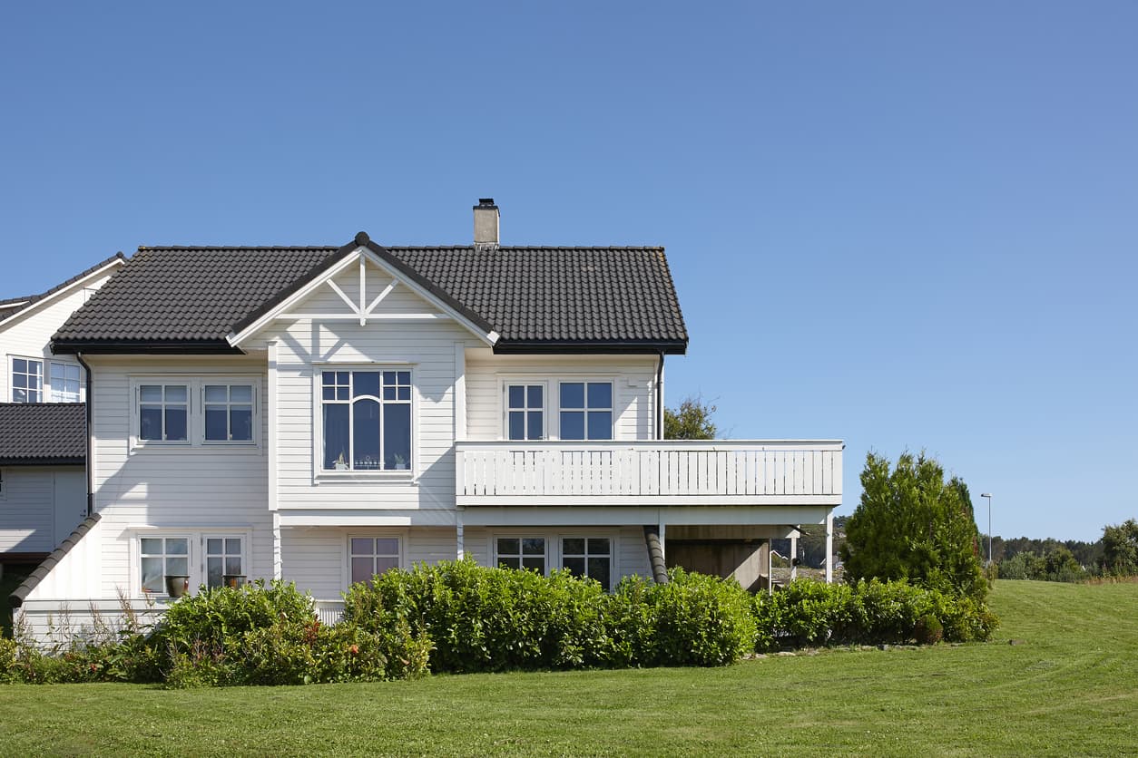 Hvit norsk enebolig med balkong og hage under blå himmel, brukt som illustrasjon i artikkel om selgers opplysningsplikt ved boligsalg.