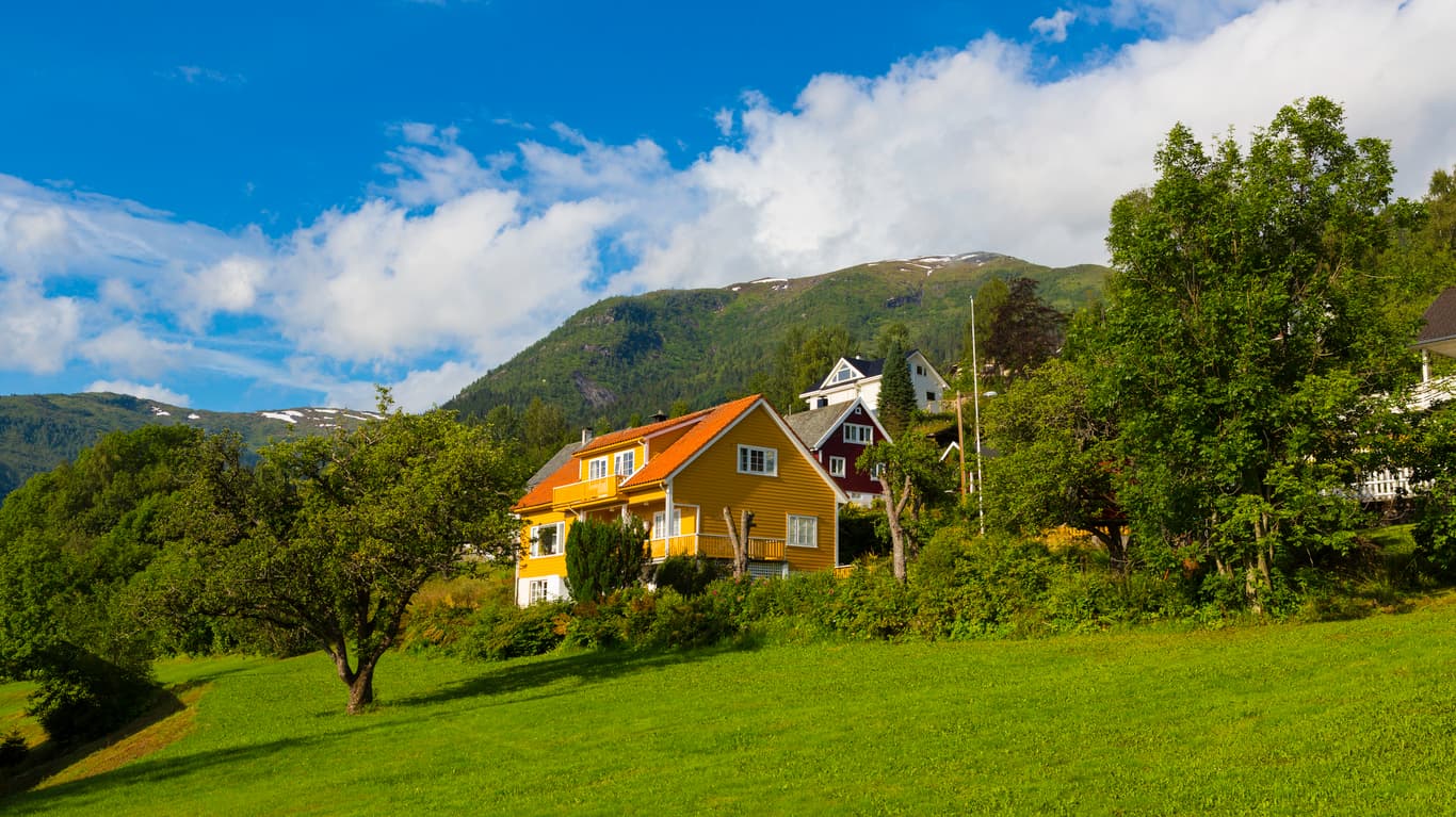 Gult trehus i grønn fjellside med frukttrær og blå himmel, brukt som illustrasjon i artikkel om avhendingsloven § 3-8. (19 ord)