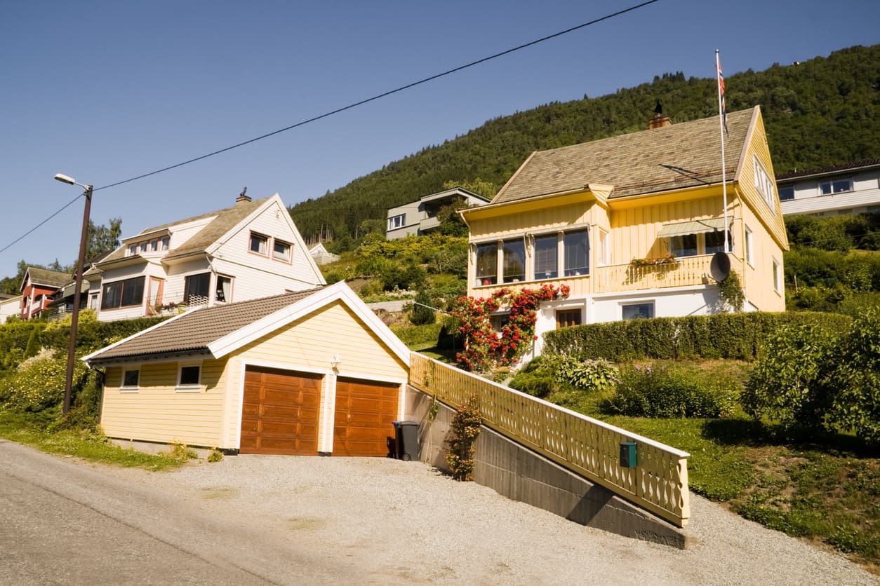 Gule trehus i en solfylt skråning med garasje og norsk flagg, brukt som illustrasjon i artikkel om dekningssalg av bolig.