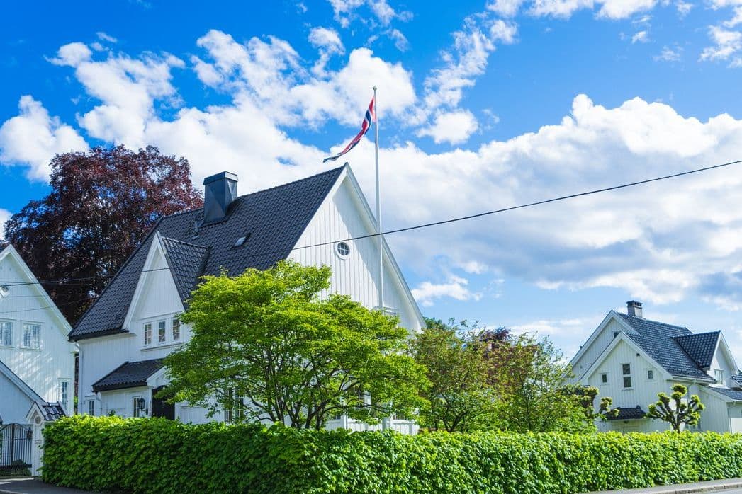 Hvit norsk enebolig med flagg og hekk under blå sommerhimmel, brukt som illustrasjon i artikkel om erstatning ved boligkjøp.