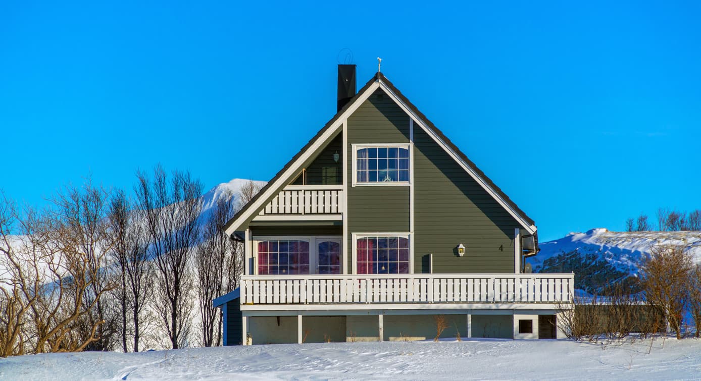 Grågrønt norsk trehus med hvit veranda omgitt av snø og fjell under blå himmel, illustrasjon til artikkel om hva avhendingsloven er.
