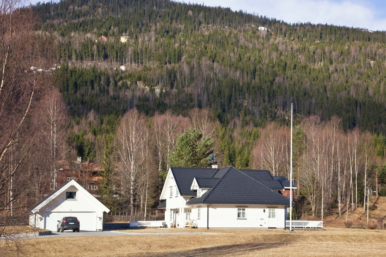 Hvit enebolig med frittstående garasje og bil i innkjørselen foran en skogkledd fjellside, brukt som illustrasjon i artikkel om hva servituttloven er.
