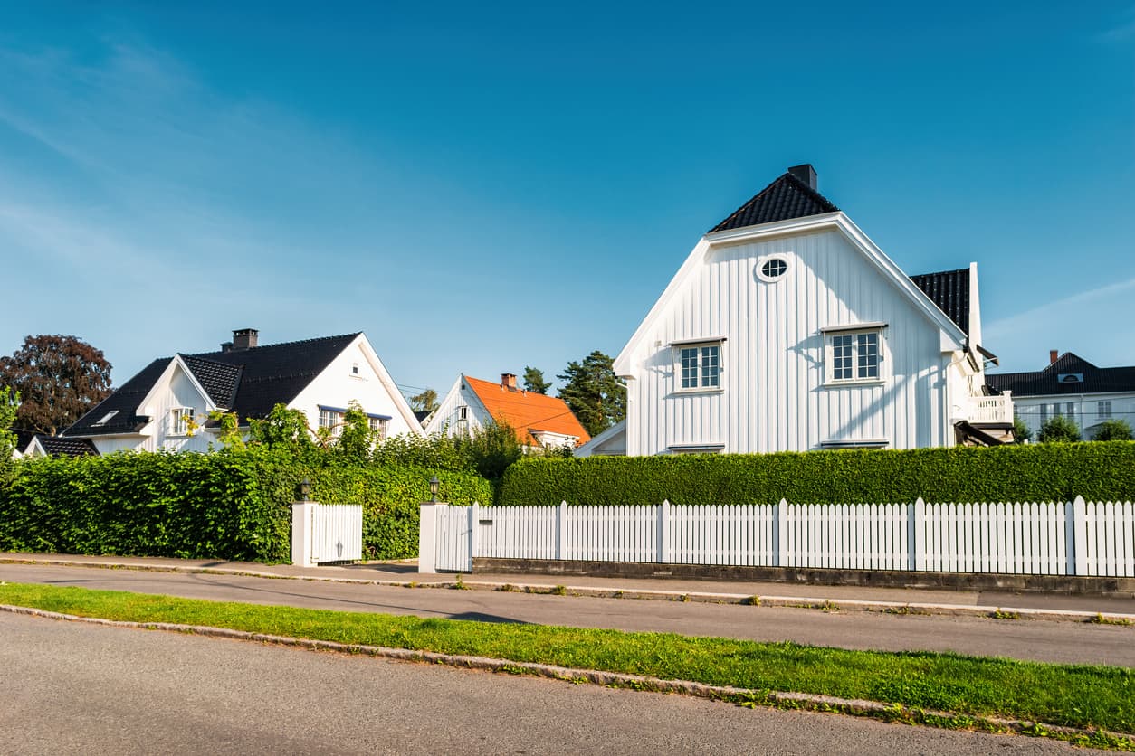 Hvitt enebolig med klippet grønn hekk og hvitt stakittgjerde mot gaten en solrik dag, brukt som illustrasjon i artikkel om naboloven.