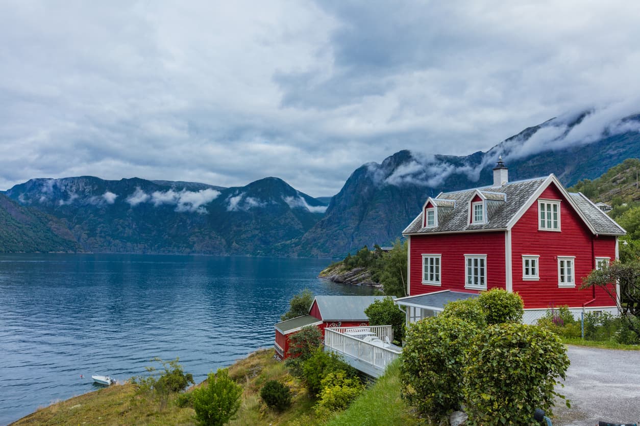 Rødt trehus med hvite vinduer ved en norsk fjord med fjell og lavt skydekke, brukt som illustrasjon i artikkel om reklamasjon på bolig.