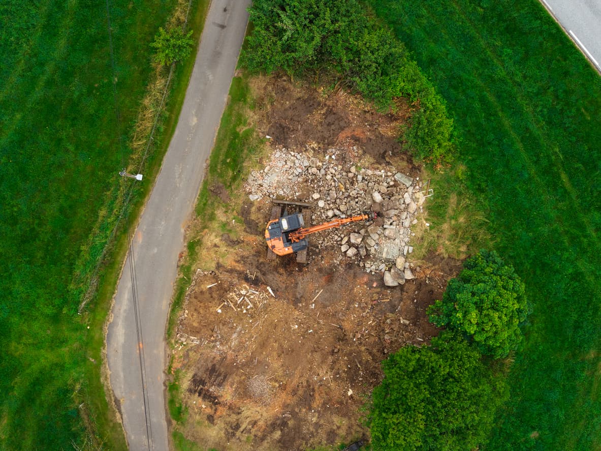 Dronebilde av en ryddet byggegrunn med oransje gravemaskin og steinmasser ved en landevei, brukt som illustrasjon i artikkel om seksjonering av tomt.