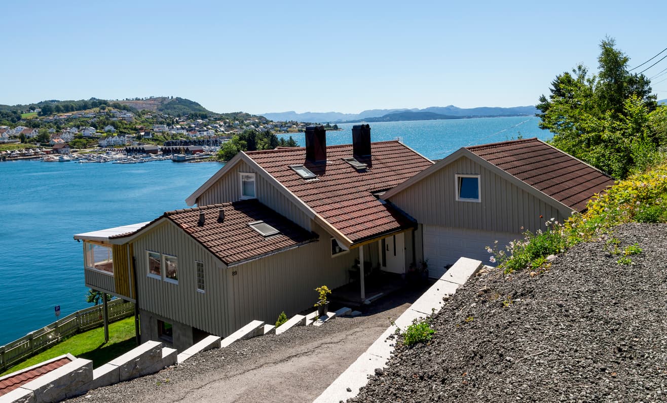 Lyst trehus med garasje på en skråtomt ved fjorden, med båthavn i bakgrunnen, brukt som illustrasjon i artikkel om tilbakeholdsrett ved boligkjøp.