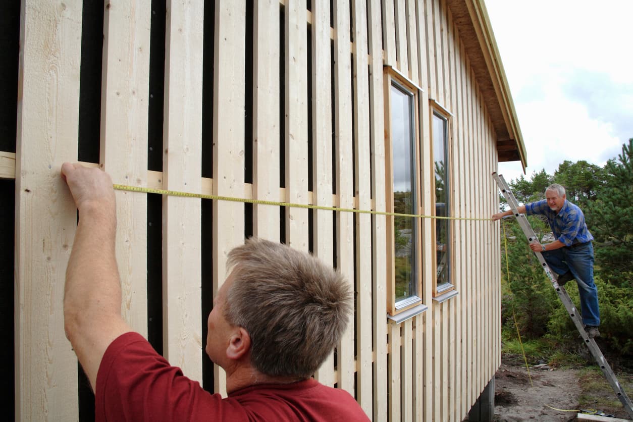 To menn måler opp og kler en trevegg med tommestokk og stige, brukt som illustrasjon i artikkel om å varsle naboer ved et byggeprosjekt.