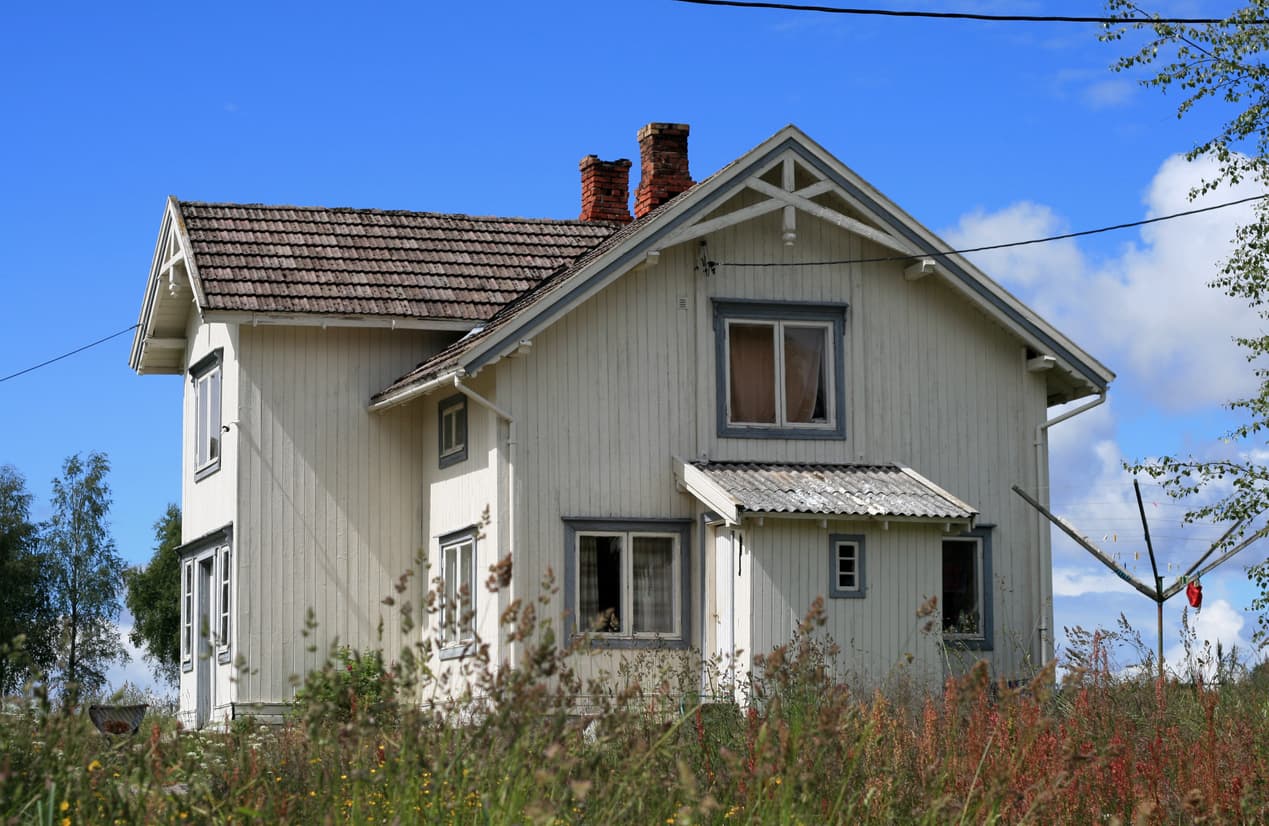 Eldre hvitt trehus med teglpiper og gresskledd tomt en sommerdag, brukt som illustrasjon i artikkel om å selge bolig til familie.