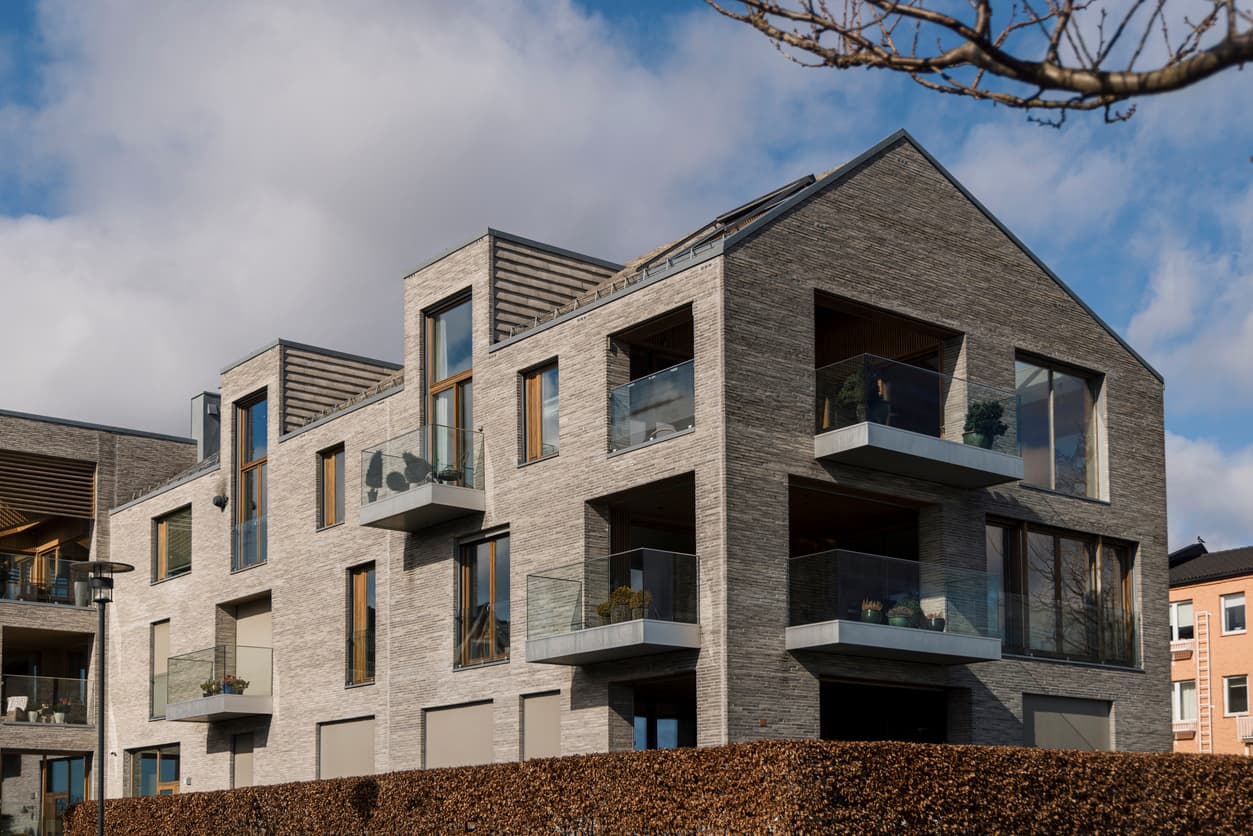Moderne leilighetsbygg i grå tegl med glassbalkonger og trevinduer bak en hekk, brukt som illustrasjon i artikkel om garanti på nybygg.