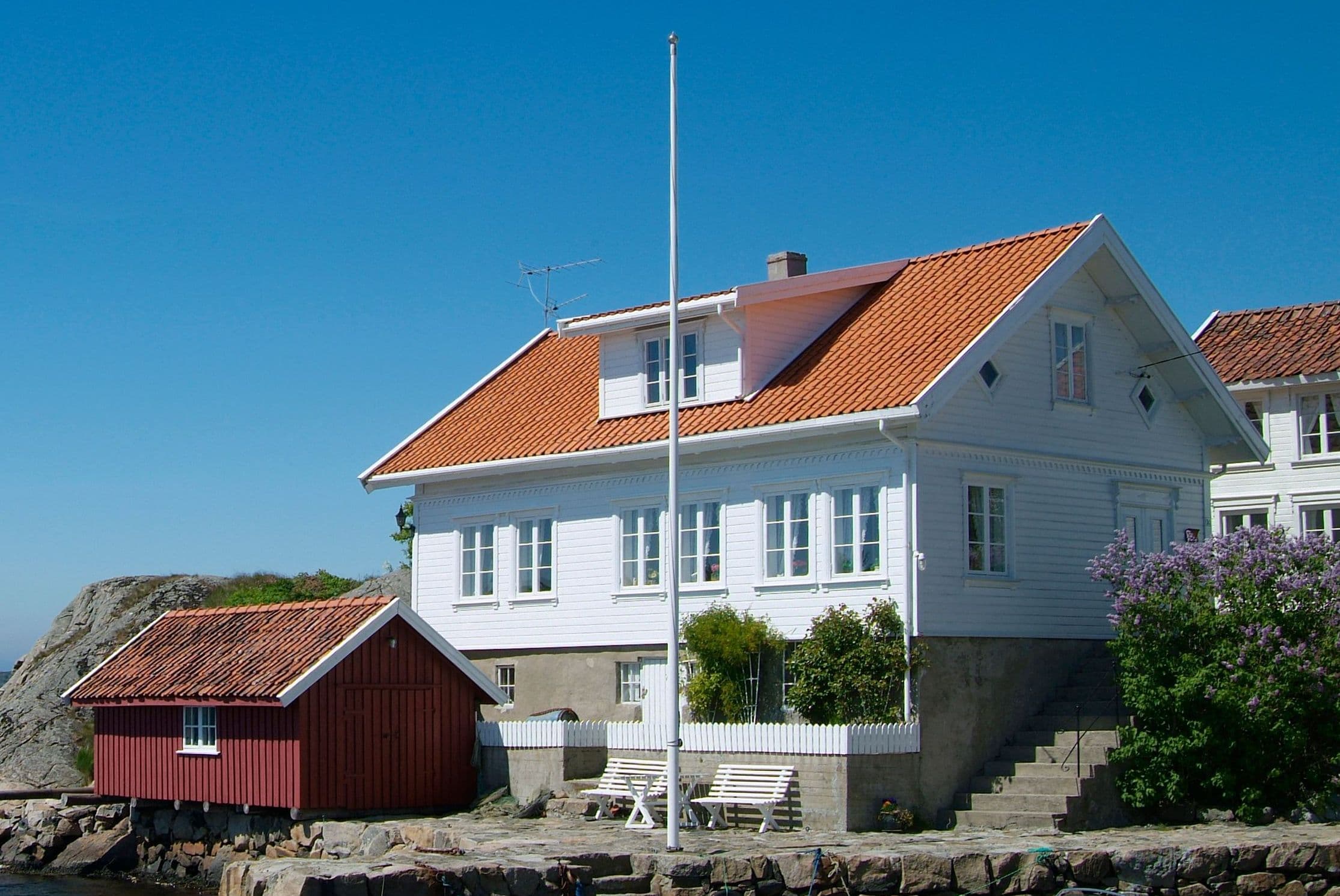 Hvitt trehus med teglstein på taket, rødt naust og flaggstang ved sjøen, brukt som illustrasjon i artikkel om klage på kjøpt eiendom.