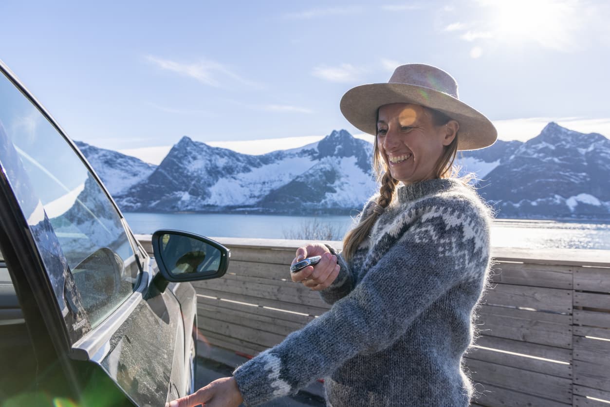 Smilende kvinne med hatt holder bilnøkkel foran åpen bildør ved fjord og snødekte fjell, brukt som illustrasjon i artikkel om forbrukerkjøpsloven om bilkjøp.