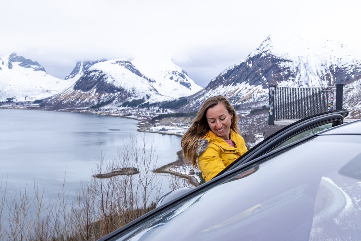 Smilende kvinne i gul jakke ved bil med snødekte fjell og fjord i bakgrunnen, brukt som illustrasjon i artikkel om kjøpers undersøkelsesplikt ved kjøp av bruktbil.