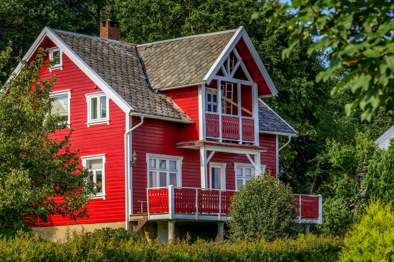 Stort rødmalt sveitserhus i tre med hvite vinduslister og verandaer omgitt av grønne busker, brukt som illustrasjon i artikkel om eiendomsoverdragelse.