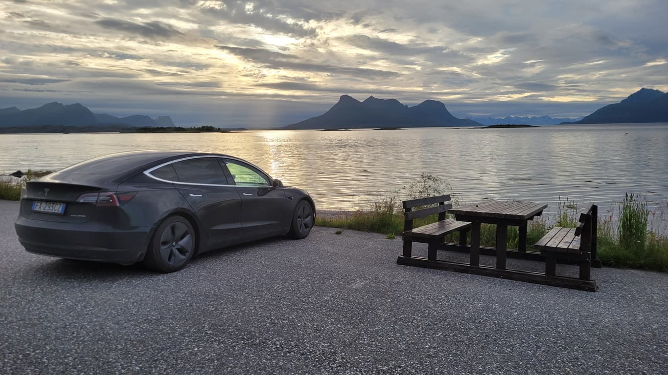 Mørk Tesla parkert ved fjord med fjell og solnedgang, brukt som illustrasjon i artikkel om når kjøpsloven og forbrukerkjøpsloven gjelder ved bilkjøp.
