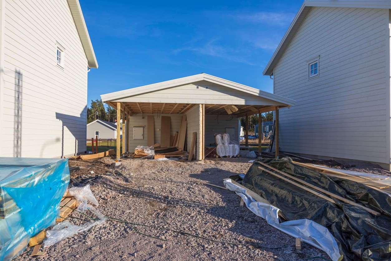 Nybygd carport mellom to hvite hus med byggematerialer på bakken, brukt som illustrasjon i artikkel om naboen har bygget på min eiendom.