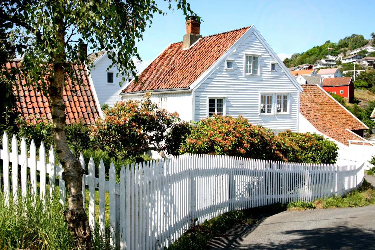 Hvitt norsk trehus med rødt tegltak og hvitt stakittgjerde om sommeren.
