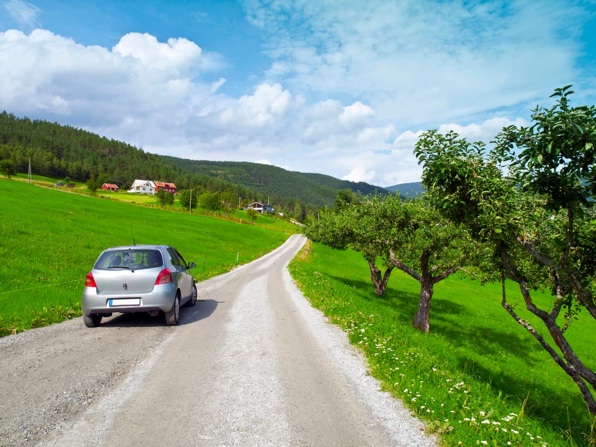 Sølvgrå bil parkert på grusvei gjennom grønne enger med hus og fjell i bakgrunnen, illustrasjon til artikkel om tinglyst veirett over annen eiendom.