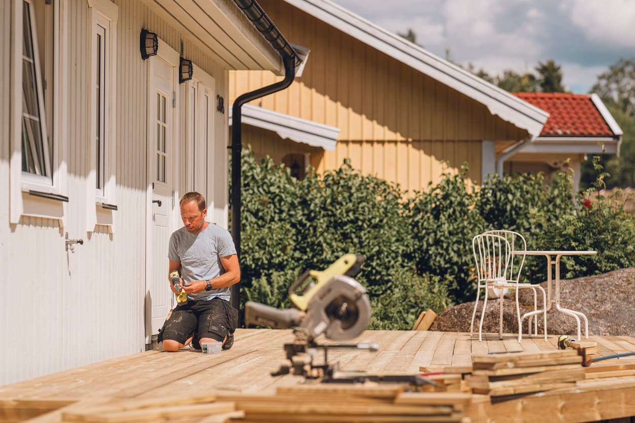 Mann monterer terrassebord ved husveggen med kappsag og verktøy, brukt som illustrasjon i en artikkel om dispensasjon fra byggegrense mot nabo.