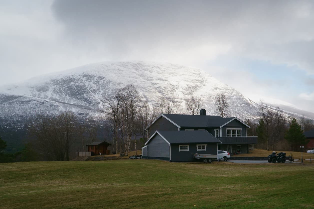 Mørk norsk enebolig på landlig tomt med snødekt fjell i bakgrunnen og åpne gressmarker, illustrasjon til artikkel om ekspropriasjonserstatning.