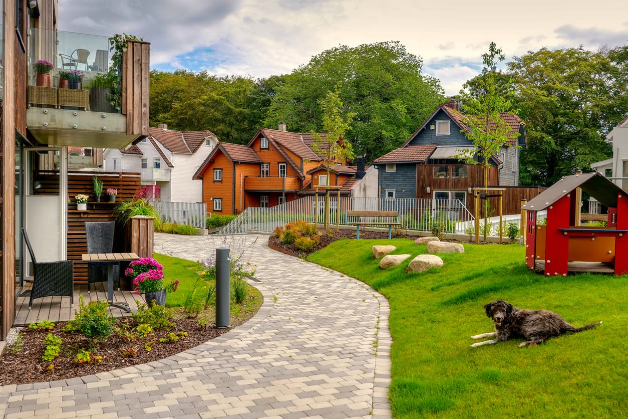 Idyllisk norsk boligfelt med fargerike trehus, brostein og blomsterbed, og en hund som ligger på plenen, brukt som illustrasjon i artikkel om naboloven § 2.