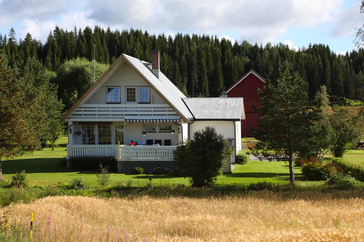Hvitt bolighus med veranda og rød låve omgitt av kornåker og skog, brukt som illustrasjon i artikkel om avhendingsloven 3-7.