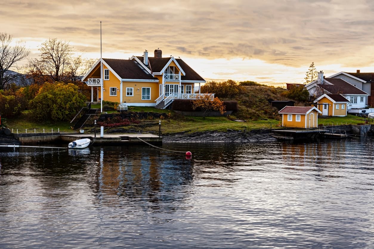 Gult trehus helt i vannkanten i solnedgang med liten båt og brygge, brukt som illustrasjon i artikkel om å bygge i strandsonen.