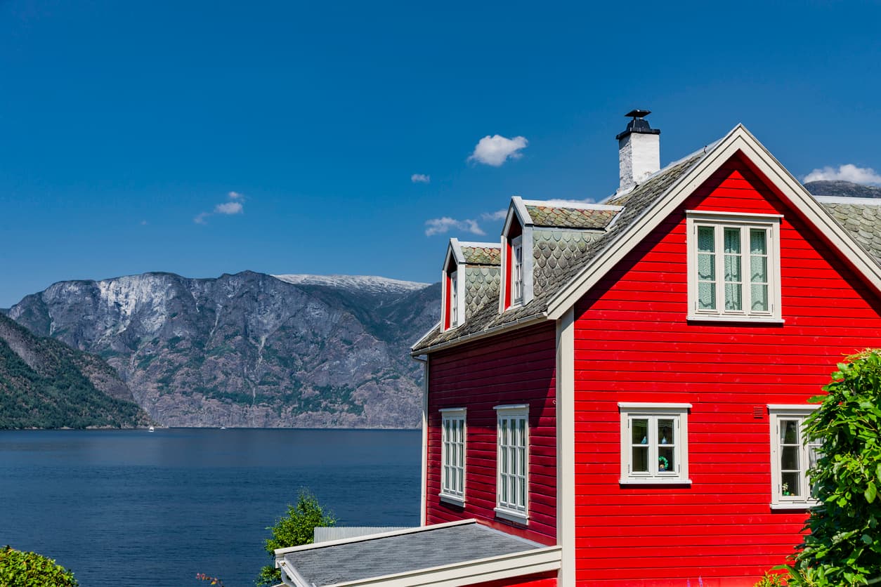 Rødt trehus med hvite vinduer ved en fjord med bratte fjell i bakgrunnen, brukt som illustrasjon i artikkel om å heve boligkjøp.
