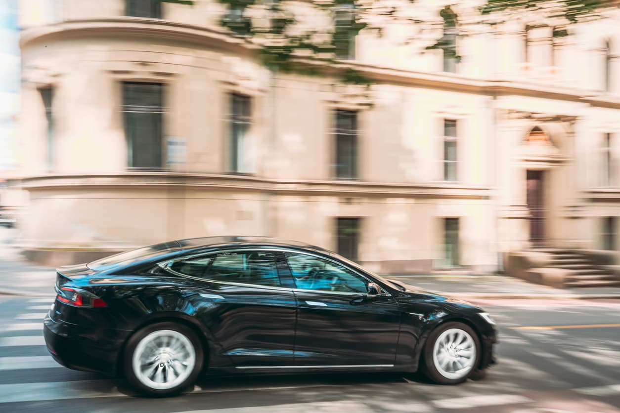 Svart Tesla i fart gjennom bygate med lys steinfasade i bakgrunnen, illustrasjon til artikkel om ansvar ved salg av bil til forhandler.