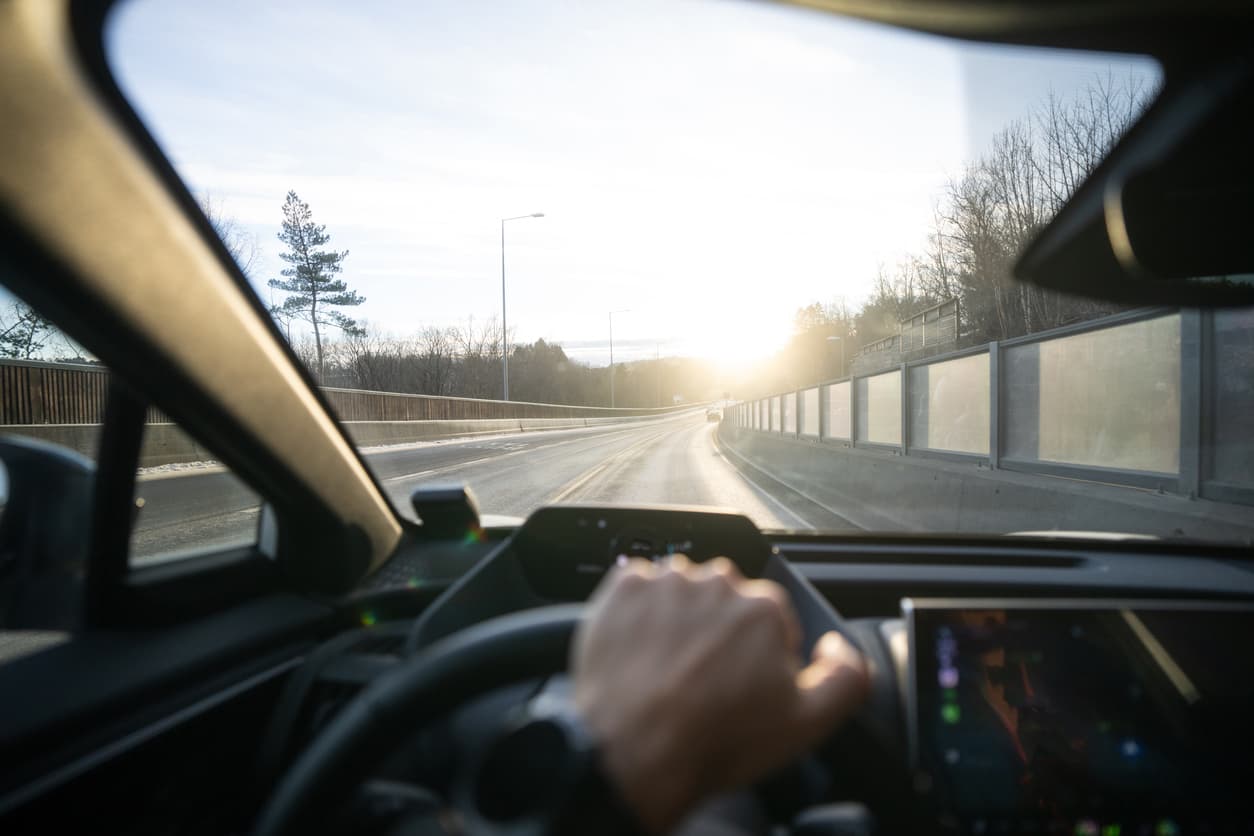 Hånd på rattet med utsikt gjennom frontruten mot vinterlig norsk vei i lavt sollys, illustrasjon til artikkel om motorhavari etter bilkjøp.