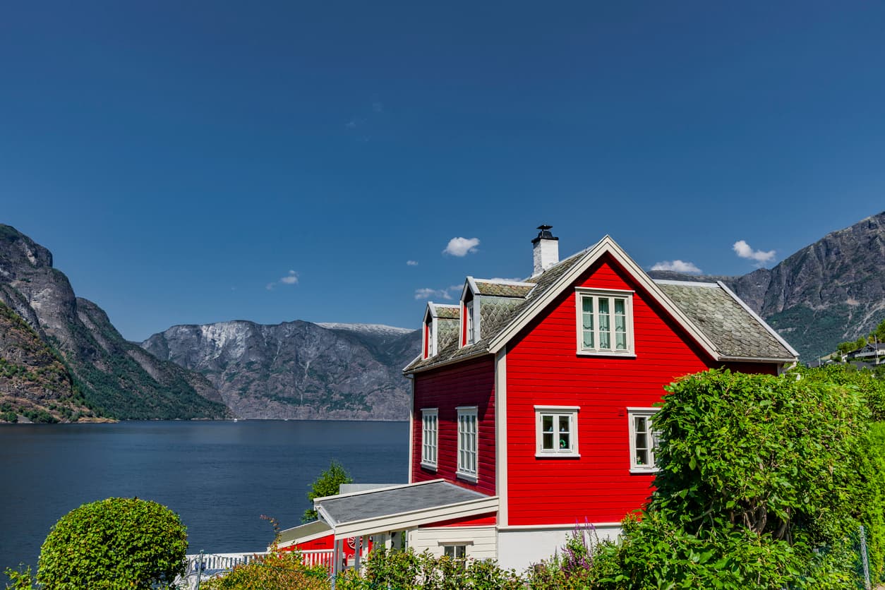 Rødt trehus ved en norsk fjord med bratte fjell i bakgrunnen, brukt som illustrasjon i artikkel om sameieloven § 15.
