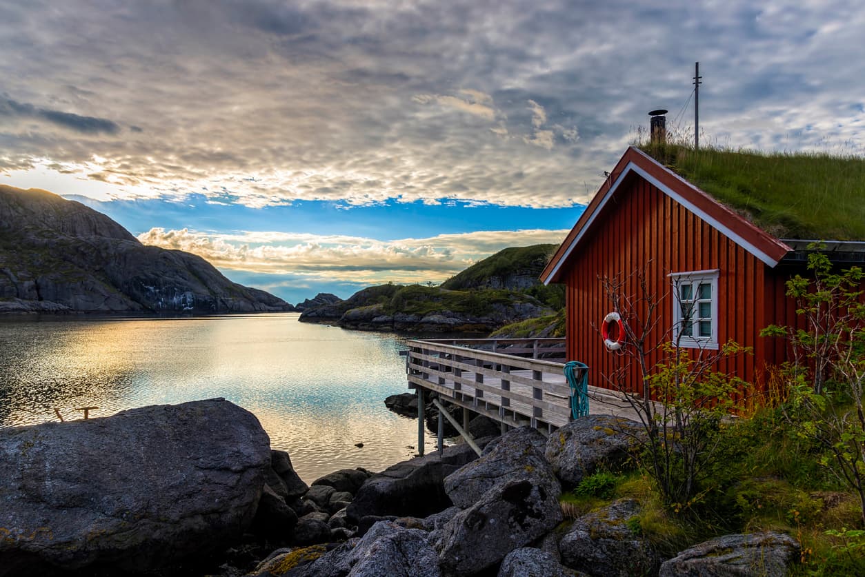 Rødmalt rorbu med gresstak og brygge ved en rolig fjord i solnedgang, brukt som illustrasjon i artikkel om avtale om sameie.
