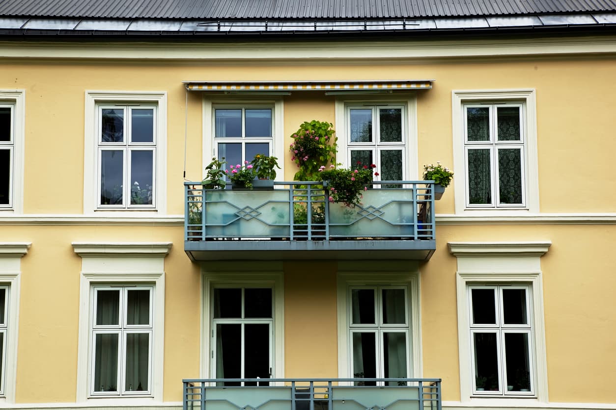 Gul bygård med utsvingt markise, blomsterkasser på rekkverket og hvite sprossevinduer, brukt som illustrasjon i artikkel om regler for balkong i sameie.