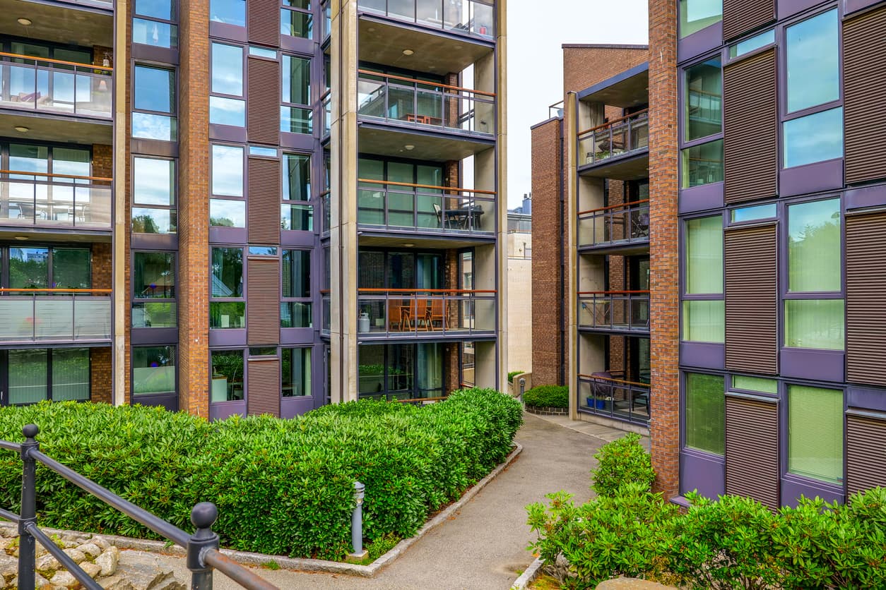 Moderne leilighetsbygg i teglstein og glass med balkonger, grønne hekker og gangvei mellom blokkene, brukt som illustrasjon i artikkel om 5 års befaring av nybygg.