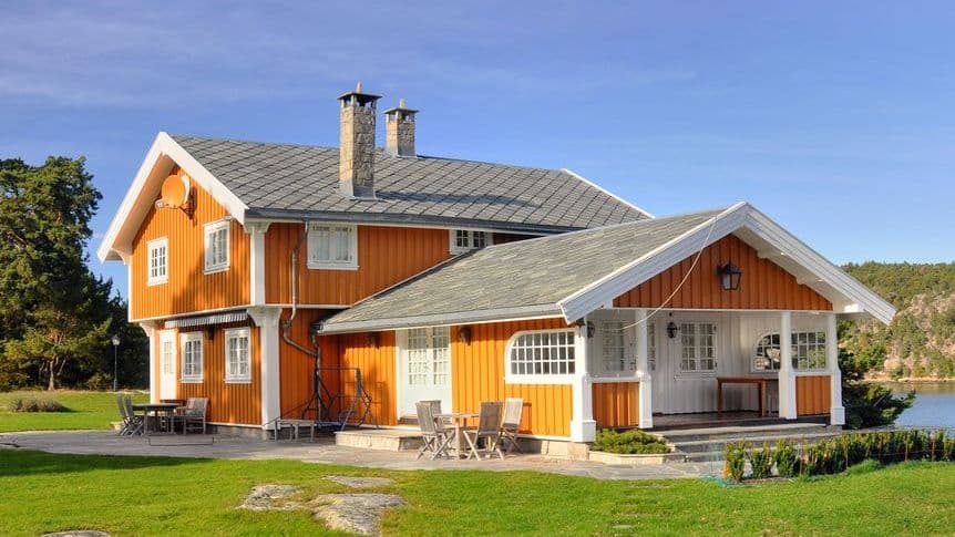 Oransje trehus med skifertak, to piper og hvite vinduer ved sjøen, illustrasjon til artikkel om hva TG2 og TG3 betyr juridisk ved boligkjøp.