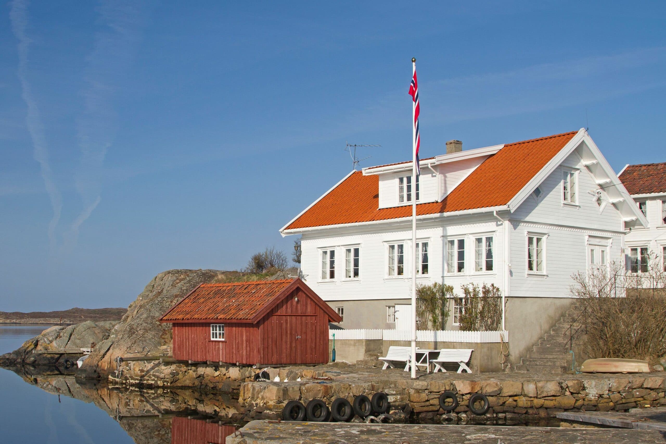 Hvitt sørlandshus med rødt tegltak, flaggstang og rødt naust ved sjøen, brukt som illustrasjon i artikkel om kjøpers undersøkelsesplikt ved boligkjøp.