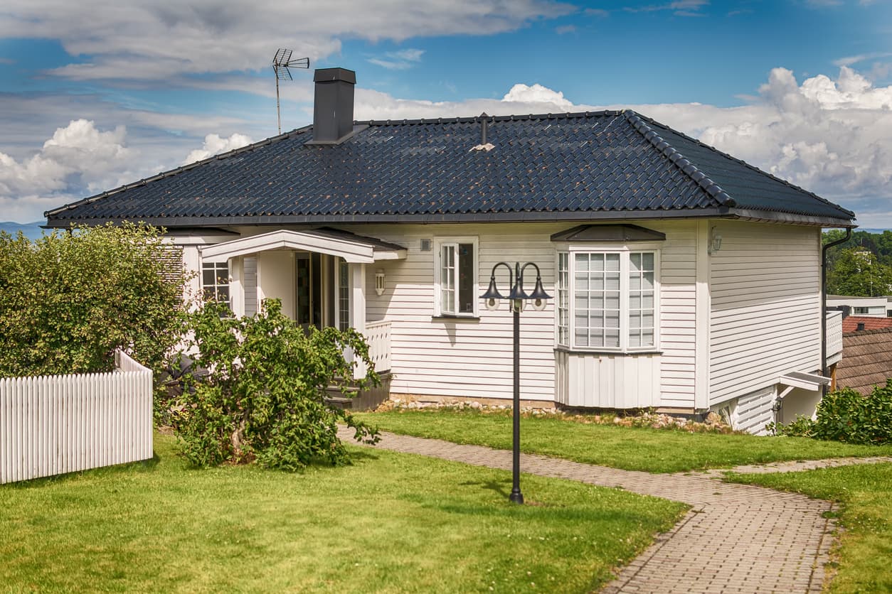 Hvitt trehus med mørkt tegltak, karnapp og stakitt i en grønn hage, brukt som illustrasjon i artikkel om reklamasjon på bolig i 5 år.