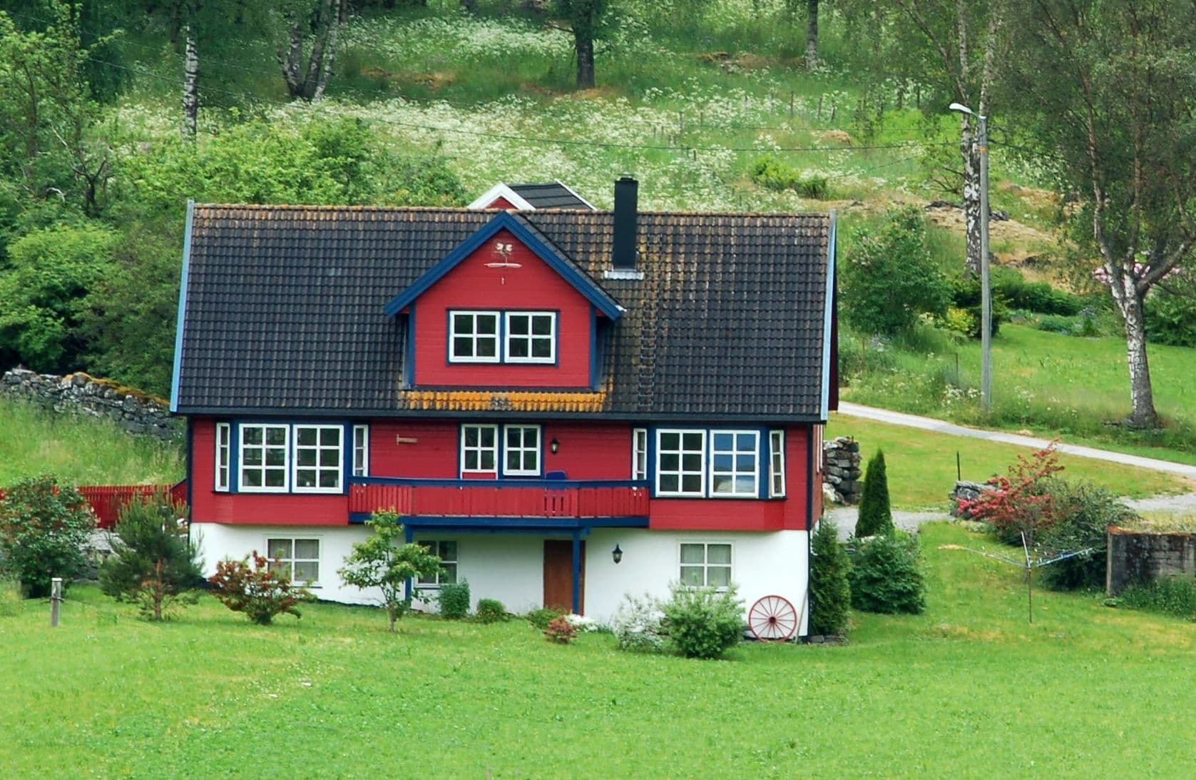 Rødt trehus med blå vinduskarmer og balkong på grønn eng, brukt som illustrasjon i artikkel om reklamasjon ved boligkjøp med mal og eksempel på reklamasjonsbrev.