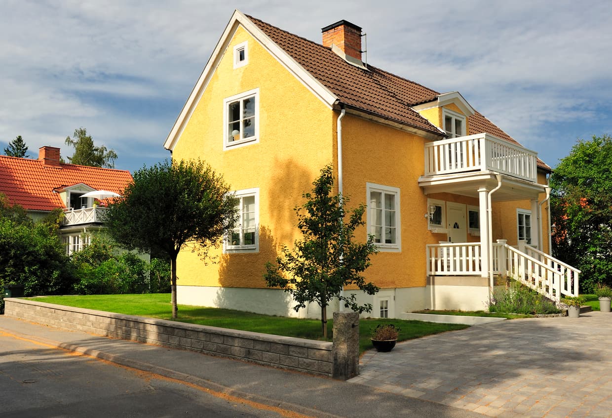 Gul pusset enebolig med hvit veranda og brunt teglsteinstak, brukt som illustrasjon i artikkel om egenandel på boligselgerforsikring.