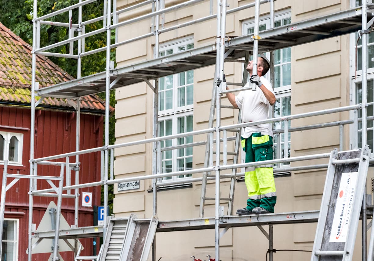 Håndverker i hvit t-skjorte og grønn-gul arbeidsbukse monterer stillas foran lys murgård i Kjøpmannsgata, brukt som illustrasjon i artikkel om innsyn i byggesak.