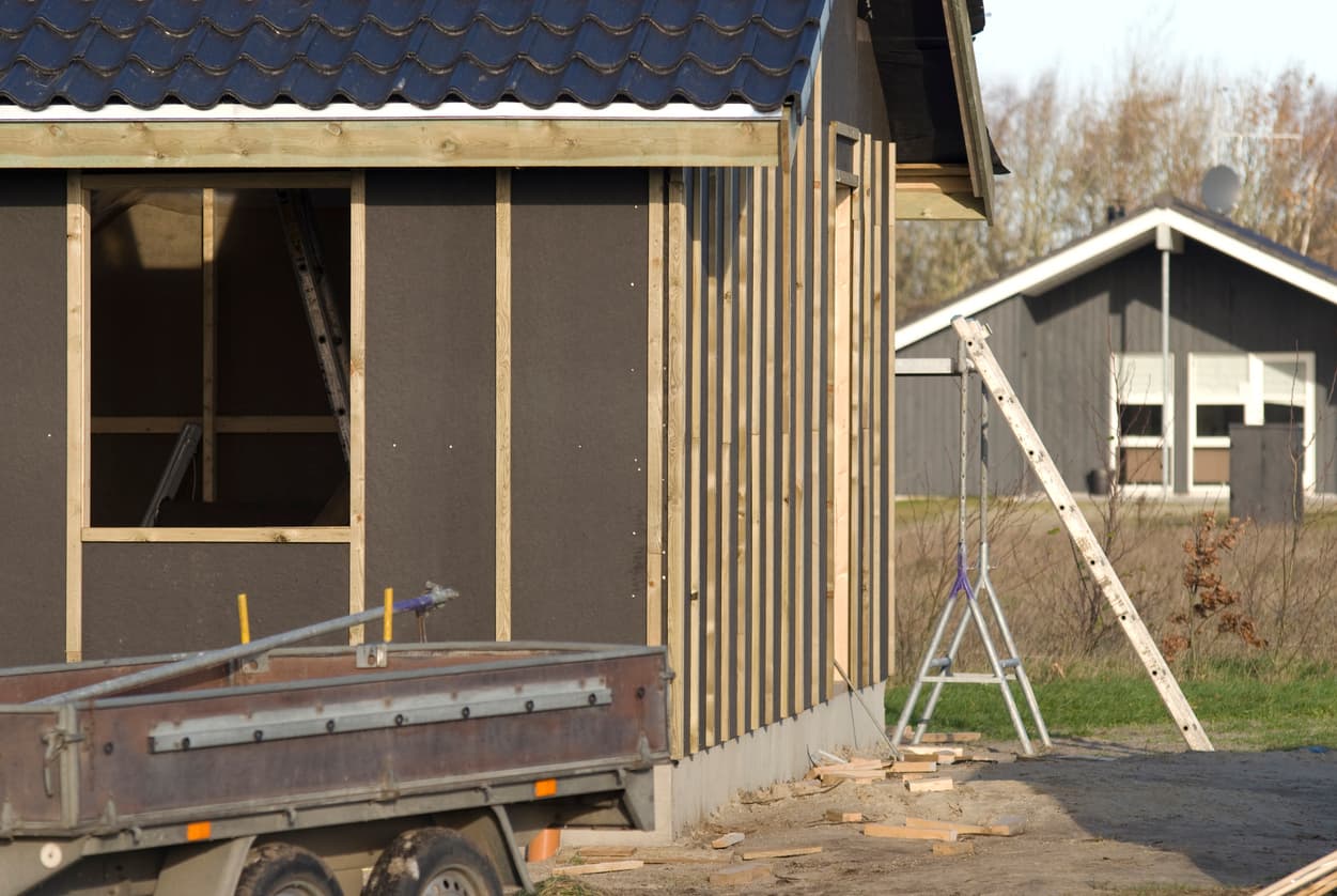 Halvferdig tilbygg med lekter, stige og tilhenger foran, tett på et grått hus i bakgrunnen, illustrasjon til sak der nabo klager på byggeprosjekt.