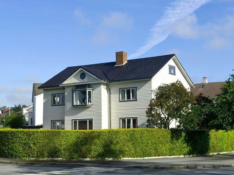 Hvitmalt enebolig med mørkt tak og høy hekk på et gatehjørne, brukt som illustrasjon i artikkel om oppgjør ved boligsalg.