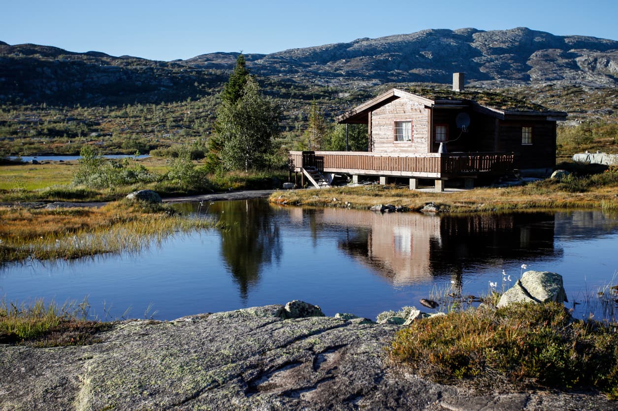 Liten tømmerhytte med torvtak og terrasse speiler seg i et fjellvann, brukt som illustrasjon i artikkel om sameieloven.