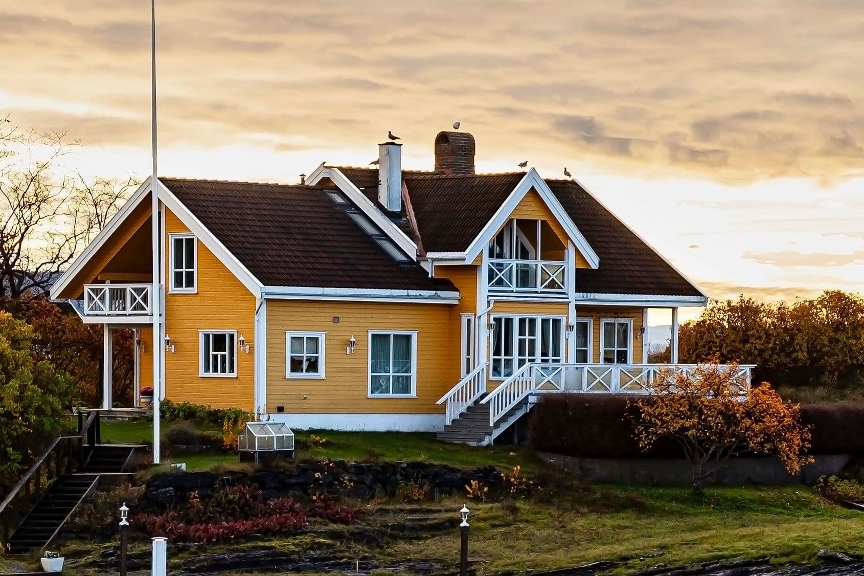 Gult trehus med hvit veranda og balkong i solnedgang, brukt som illustrasjon i artikkel om fritt advokatvalg ved boligkjøperforsikring.