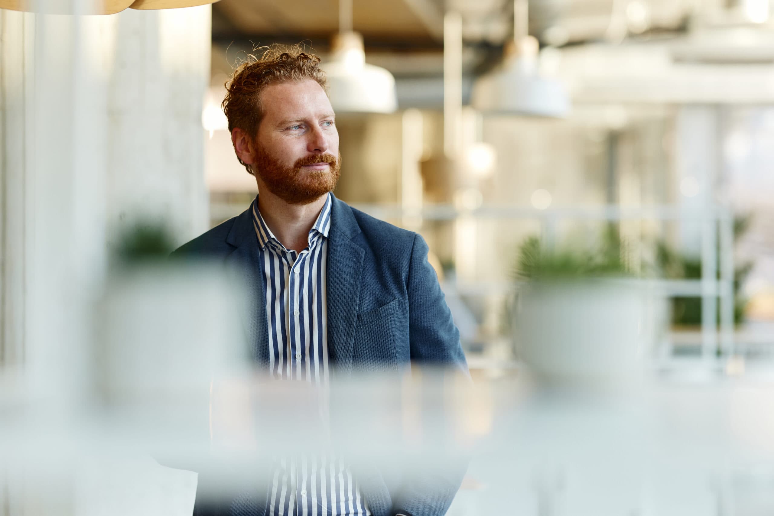 Mann med rødt skjegg og blå blazer ser ut av vinduet i et lyst kontor, brukt som illustrasjon i artikkel om arbeidsmiljøloven og overtid.